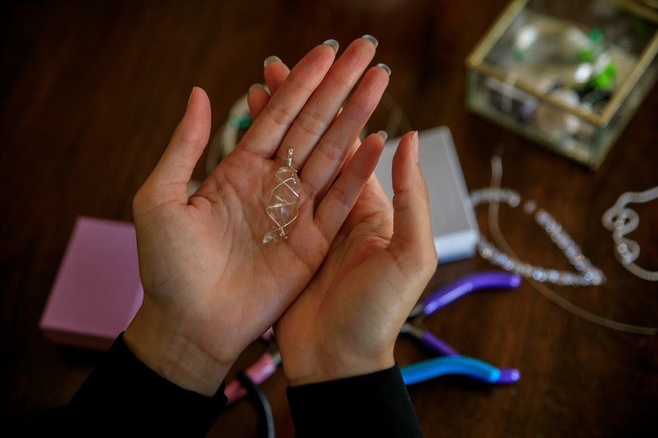 home-hero-1 Close-up of hands presenting a handmade silver wire pendant with jewelry tools on a wooden table.